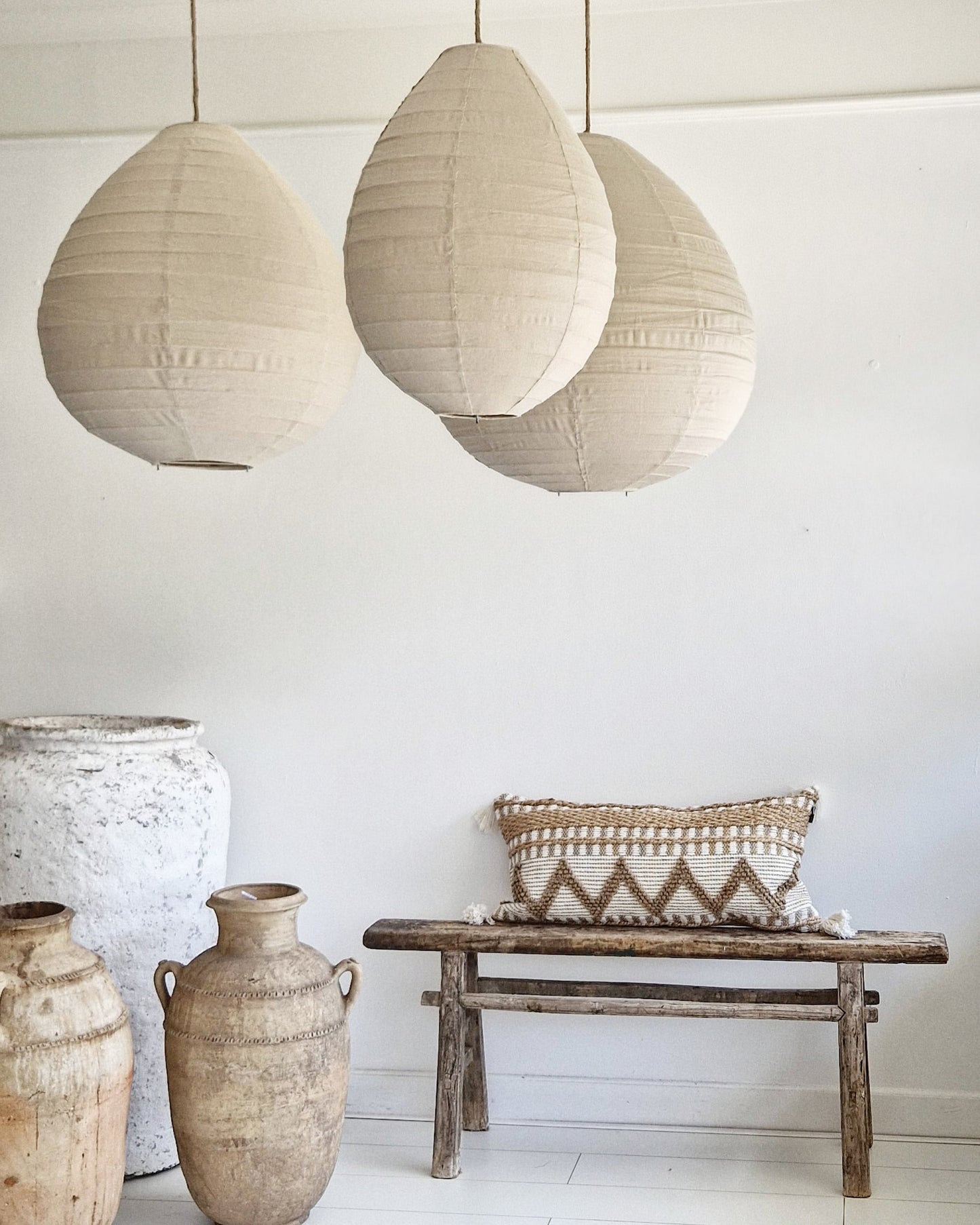 Rustgevend wit interieur met linnen lampion hanglampen in de kleur Sand, die een zachte, natuurlijke sfeer creëren. De Marokkaanse aardewerken kruiken en het oude houten Chinese bankje met prachtig langwerpig kussen in naturel kleuren versterken de minimalistische en serene uitstraling van de ruimte.