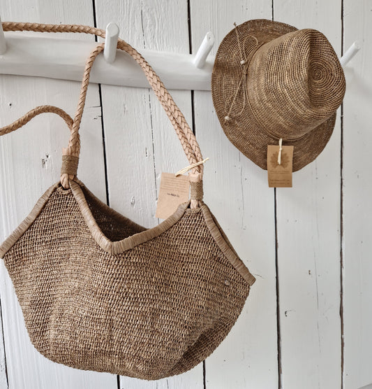 Aina M Bag van Made in Mada met hoed aan witte wandhaak, handgeweven raffiatas met gevlochten leren hengsels in warme Tea-tint