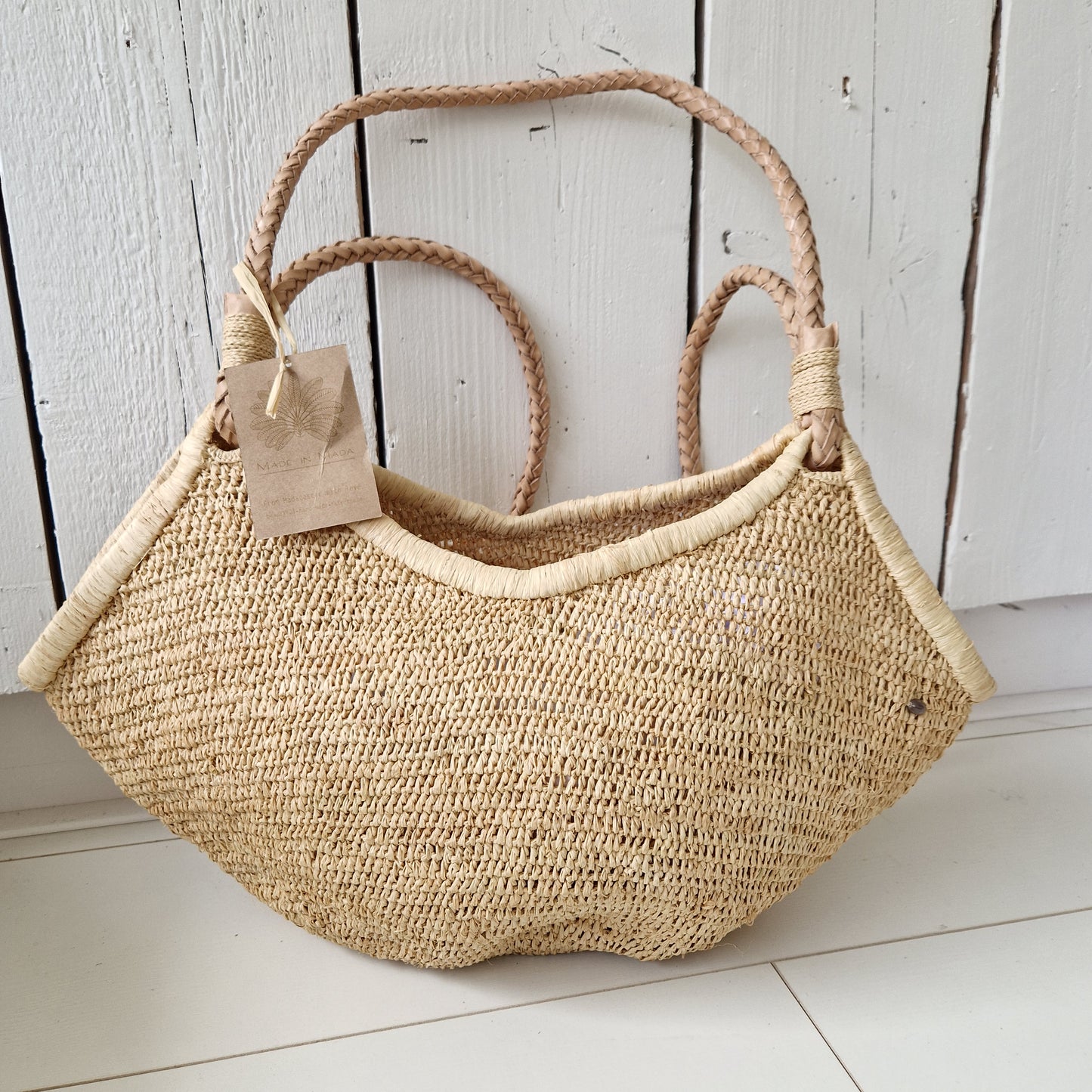 Aina M Bag Naturel rustend op witte vloer, handgeweven raffiatas met lichte structuur en leren hengsels
