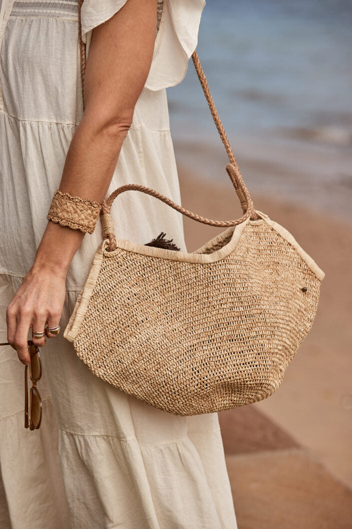 Aina M Bag van Made in Mada gedragen op het strand, licht raffiaweefsel met leren hengsel bij een luchtige jurk