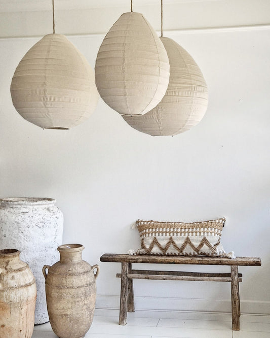 Rustgevend wit interieur met linnen lampion hanglampen in de kleur Sand, die een zachte, natuurlijke sfeer creëren. De Marokkaanse aardewerken kruiken en het oude houten Chinese bankje met prachtig langwerpig kussen in naturel kleuren versterken de minimalistische en serene uitstraling van de ruimte.